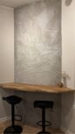 Textured light grey plaster wall behind a bar counter with a timber top and two black stools
